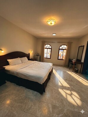 A hotel room with a large bed, two bedside tables, a desk with a chair, and two windows with curtains.