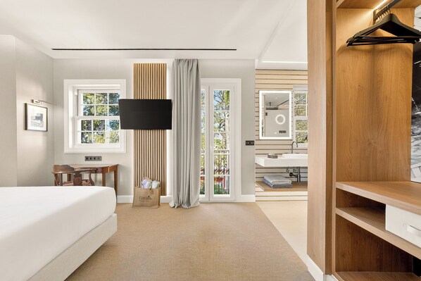 A modern hotel room with a bed, a desk, a TV, a bathroom with a sink and mirror, and a view of the outdoors.