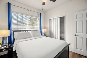 Ett hotellrum med en säng, sänglampor, ett fönster med persienner, en dörr och en takfläkt.