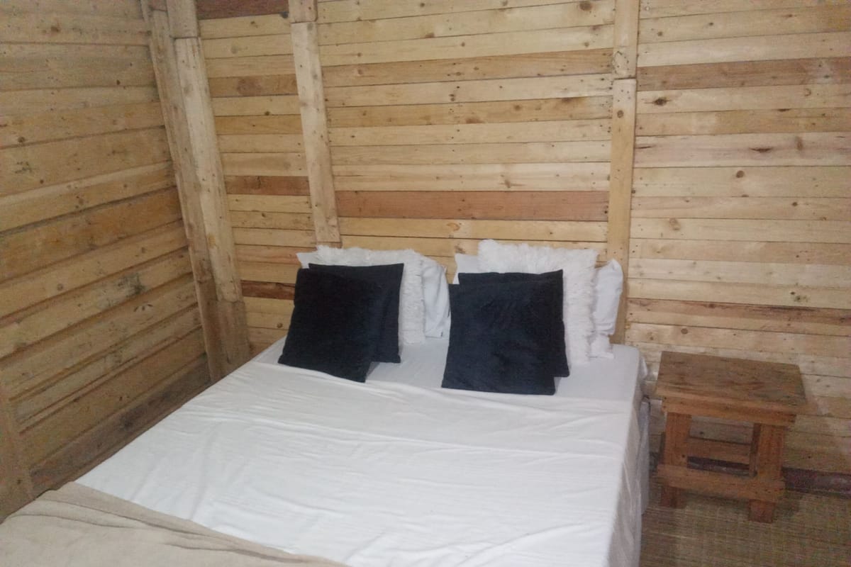 Una habitación tipo cabaña de madera con una cama, almohadas y un taburete de madera.