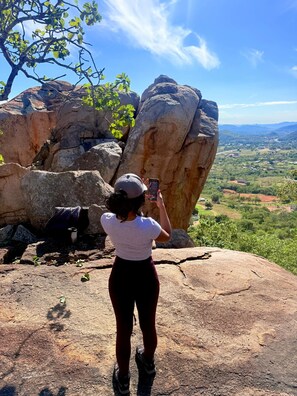 A person taking a photo with a smartphone, standing on a rocky surface with large boulders and a tree in the background.