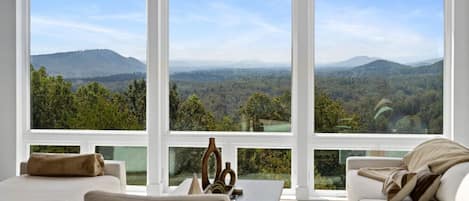 Habitación con un ventanal grande que da a un paisaje boscoso y montañas. Hay un sofá blanco y una mesita con un florero.