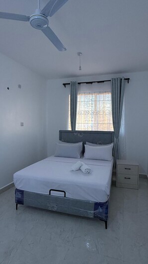 Un dormitorio con cama, ventilador de techo, ventana con cortinas y mesita de noche.