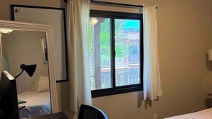 Una habitación con cama, escritorio, silla, ventana con cortinas y vistas a edificios.