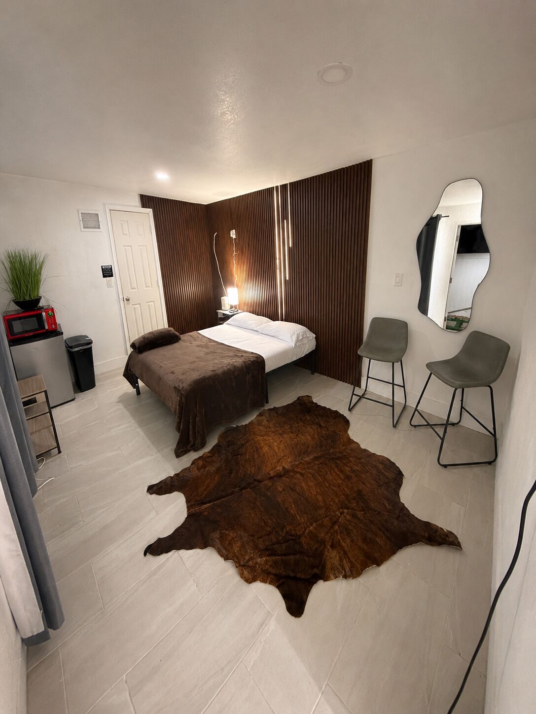 A bedroom with a bed, two chairs, a mirror, a potted plant, a microwave, and a wooden panel wall.