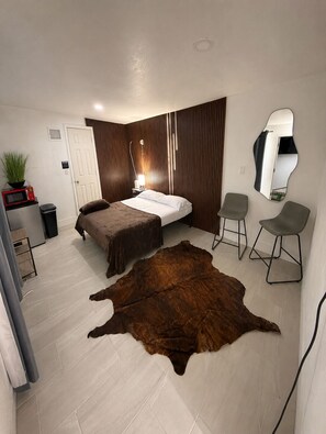 Une chambre à coucher comprenant un lit, deux chaises, un miroir, une plante en pot, un four à micro-ondes et un mur recouvert de panneaux de bois.
