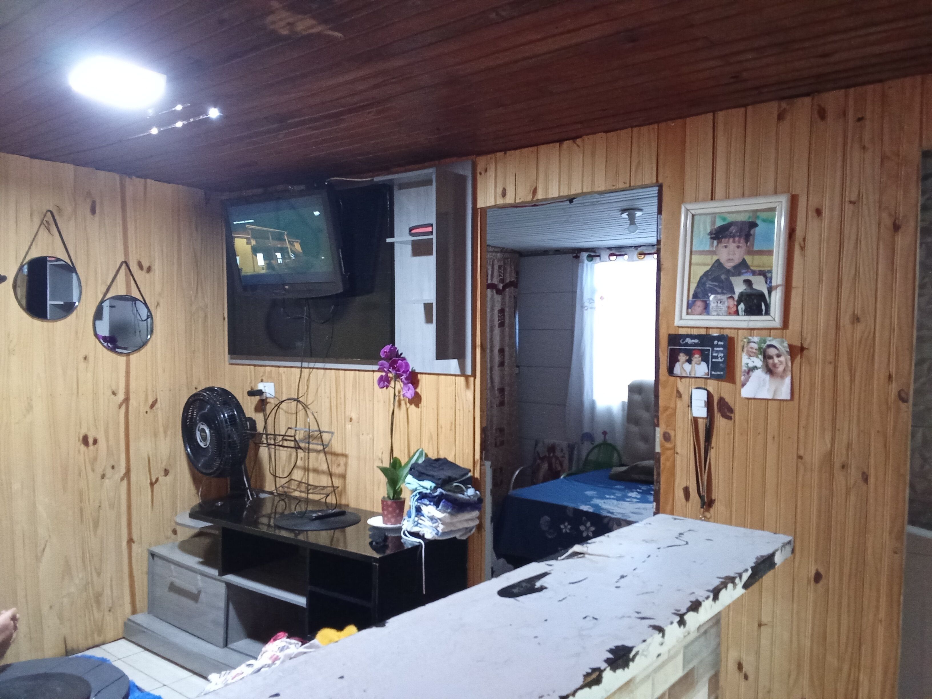 Quarto com paredes de madeira, televisão, escrivaninha com computador, cadeira e cama.