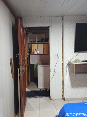 Ein Zimmer mit Holztür, einer Waschmaschine, einem Regal mit Gegenständen und einem an der Wand befestigten Fernseher.