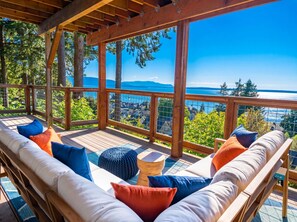 Une terrasse en bois avec un canapé et des coussins, offrant une vue panoramique sur une étendue d’eau et un paysage lointain.