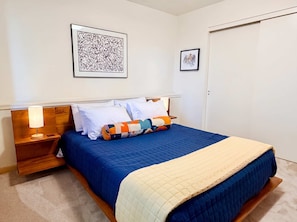 Una cama bien hecha con un edredón azul y una manta color beige, dos lámparas de noche y una obra de arte abstracta enmarcada sobre la cama.