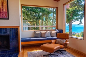 A cozy seating area with a sofa and ottoman, a fireplace, and a large window offering a view of trees and a body of water. - Luxury Treehouse Retreat | Hot Tub + Bay Views (Bellingham)