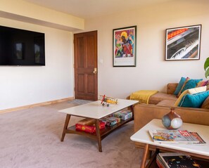 A living room with a flat-screen TV, a wooden door, a coffee table with magazines, a sofa with cushions, and two framed pictures on the wall. - Luxury Treehouse Retreat | Hot Tub + Bay Views (Bellingham)