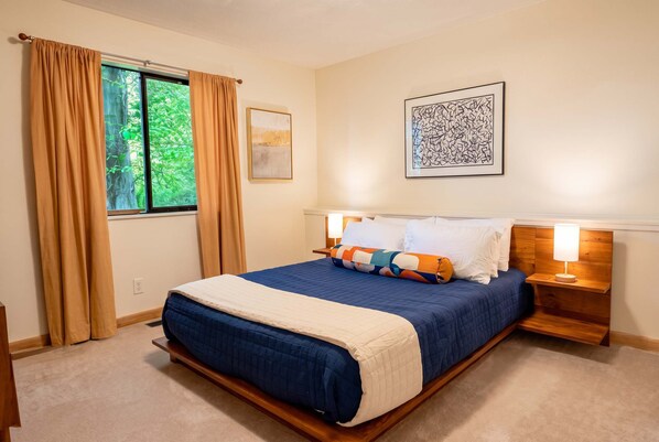 A bedroom with a large bed, two bedside tables, a window with curtains, and two wall-mounted artworks.