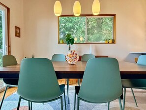 A dining area with a wooden table, green chairs, pendant lights, and a window with a view of trees.