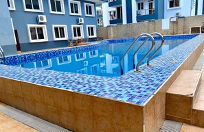 A swimming pool with blue mosaic tiles, surrounded by a building with multiple windows and air conditioning units. - Lovely 3-bedroom apartment near the beach (Dar es Salaam)