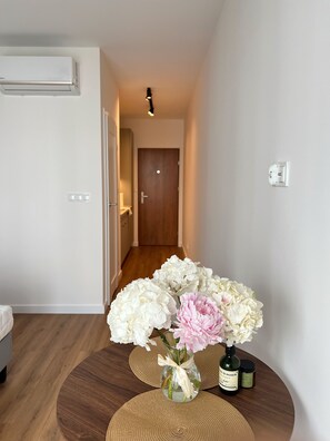 Un pasillo con una puerta de madera al final, una unidad de aire acondicionado blanca en la pared, y una mesa con un jarrón de flores y una botella.