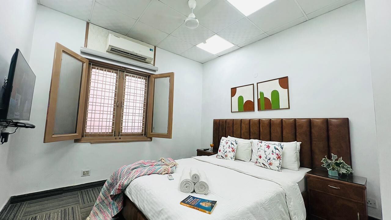 A hotel room with a bed, a bedside table, a nightstand, a TV, an air conditioning unit, and a window with a view.
