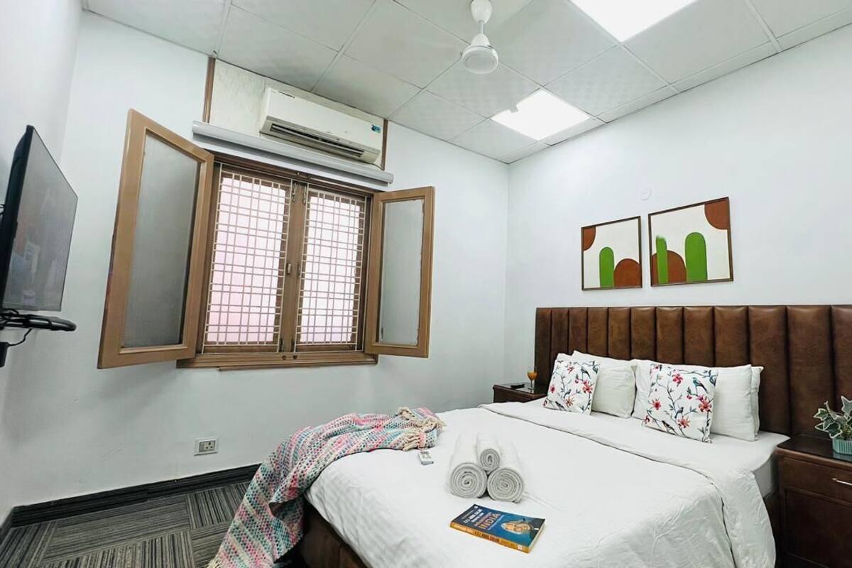 A hotel room with a bed, a bedside table, a nightstand, a TV, an air conditioning unit, and a window with a view.