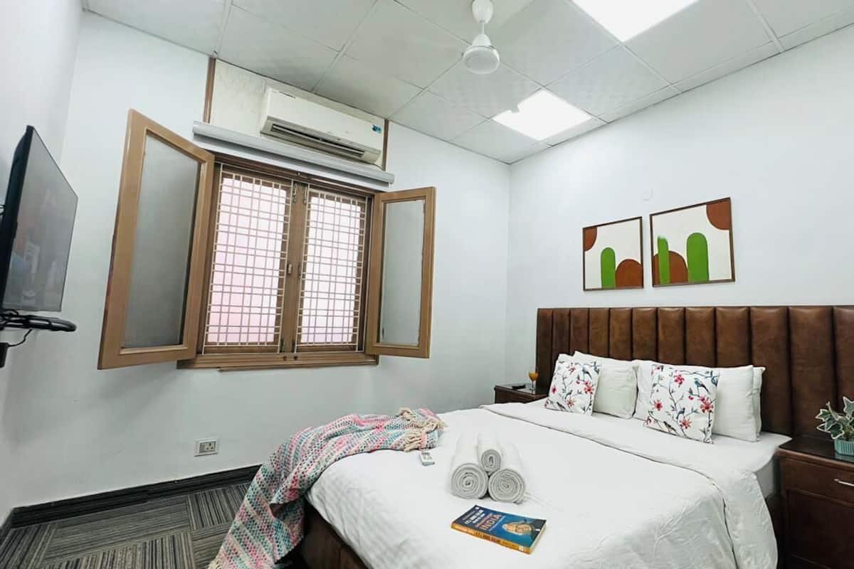 A hotel room with a bed, a bedside table, a nightstand, a TV, an air conditioning unit, and a window with a view.