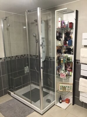 Et brusebad med glasvægge, flisegulv, en glashylde med toiletartikler og et hvidt håndklædestativ.