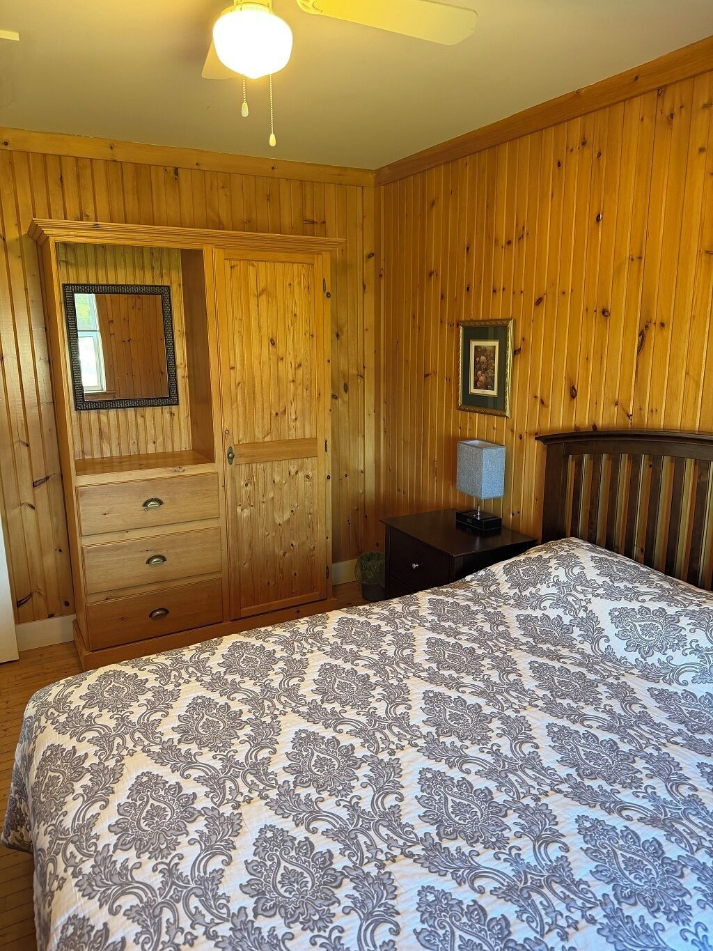 Een slaapkamer met houten lambrisering, een bed met beddengoed met een patroon, een dressoir, een spiegel, een ingelijst schilderij en een nachtkastje.