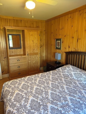 Un dormitorio con paneles de madera, una cama con ropa de cama estampada, un tocador, un espejo, un cuadro enmarcado y una mesita de noche.