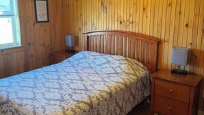 Un dormitorio con pared de paneles de madera, una cama con colcha estampada, un ventilador de techo y una mesita de noche con lámpara.