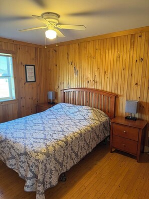 A bedroom with a wooden paneled wall, a bed with a patterned bedspread, a ceiling fan, and a nightstand with a lamp. - Captain's Den Cottage (Brule Shore)