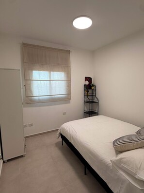 A bedroom with a bed, a window with a blind, a bedside table with a lamp, and a door. - K-Home 3 Bedroom Karme Gat - Kiriat Gat (קריית גת)