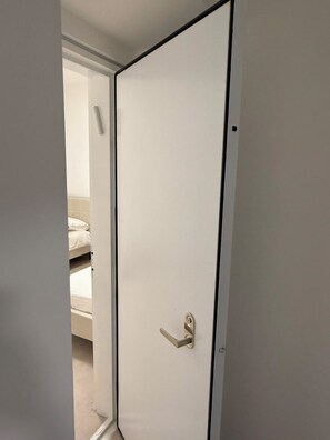 A closed white door with a silver handle, a visible gap at the bottom, and a partial view of a bed with white bedding in the background. - K-Home 3 Bedroom Karme Gat - Kiriat Gat (קריית גת)