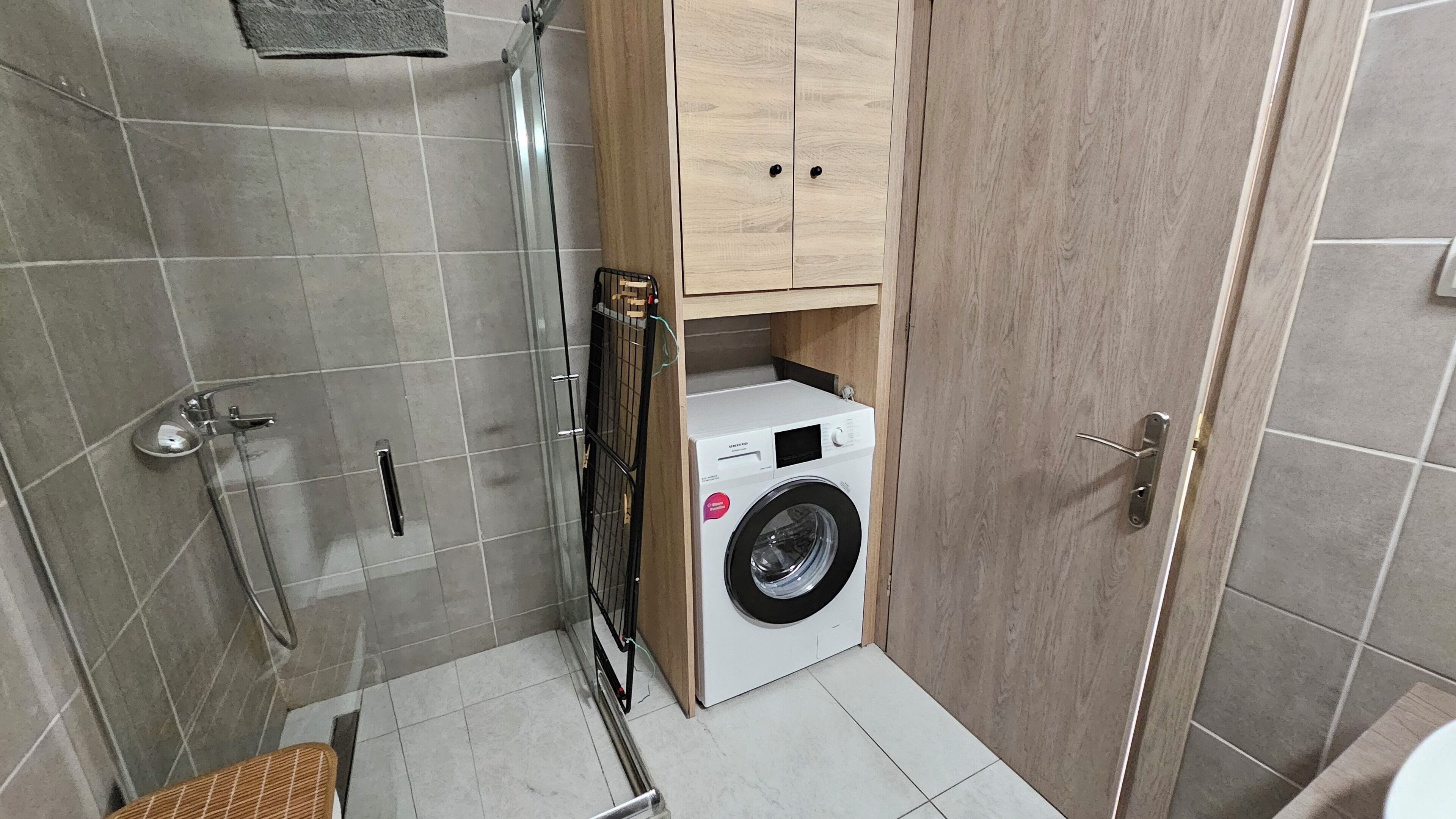 Een kleine badkamer met een douche, een wasmachine en houten kastjes.