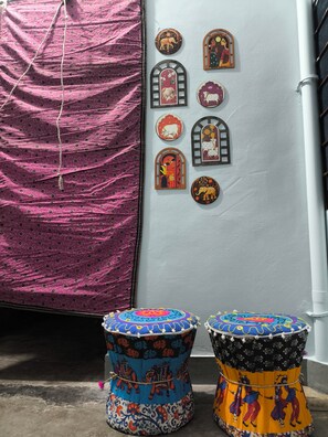 A room with a pink patterned fabric draped over a chair, a white wall with decorative wooden cutouts, and two colorful stools with intricate designs. - 2kms  to hawamahal homestay in jaipur (Jaipur)
