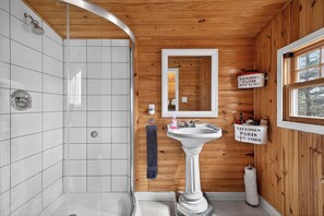 A bathroom with a shower, a pedestal sink, a mirror, and wooden walls.