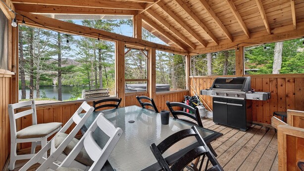 A wooden deck with a glass table and chairs, a grill, and a view of a lake.