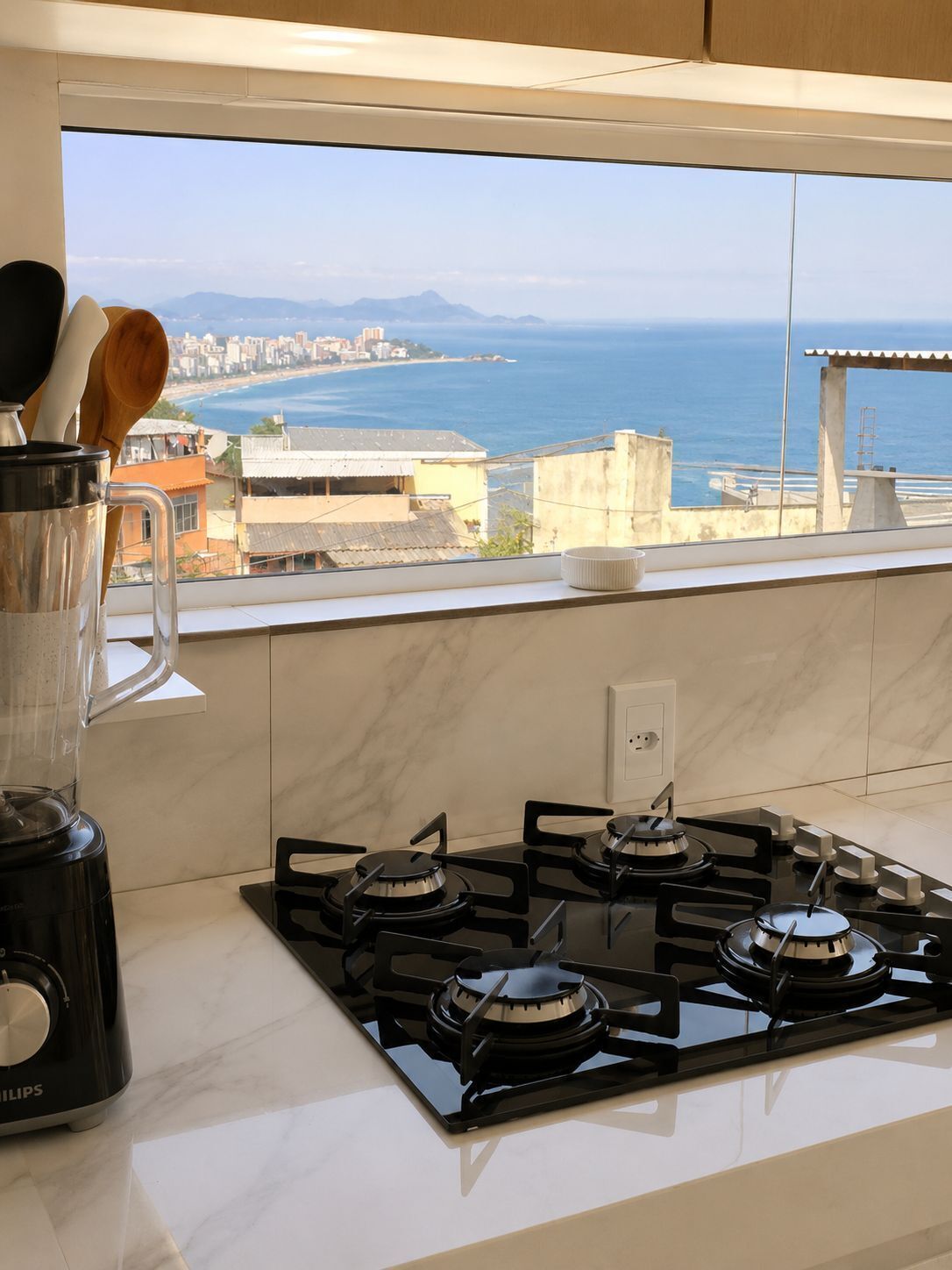 A kitchen with a gas stove, a blender, and a window with a view of the sea and buildings.