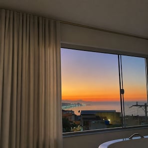 A room with a large window, a curtain, and a view of a cityscape at sunset. - Ap Favela Chic, very cozy. With a hot tub and barbecue, 700 meters from the beach. (Rio de Janeiro)