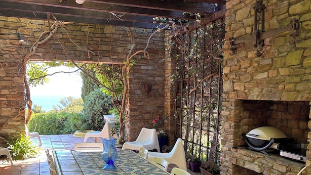 Eine überdachte Terrasse mit Steinwand, Fliesenboden, Tisch mit Stühlen und Grillbereich.