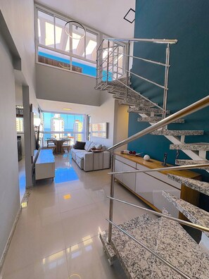 Ein modernes Interieur mit hoher Decke, einer Treppe mit Metallgeländern, einem Essbereich mit langem Tisch und Blick auf die Stadt durch große Fenster.