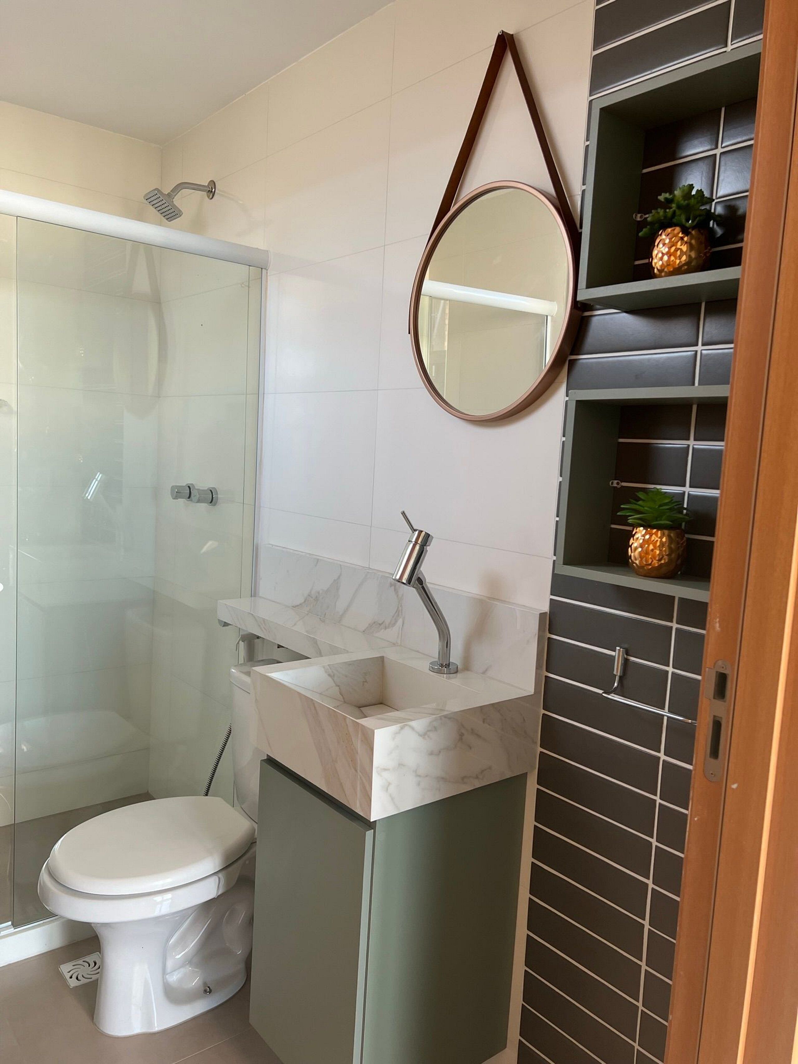 A bathroom with a glass-enclosed shower, a toilet, a sink with a marble countertop, a round mirror, and a shelf with decorative items.
