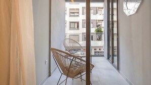 Un balcó estret amb una cadira de vimet, una planta en test i una decoració mural geomètrica.