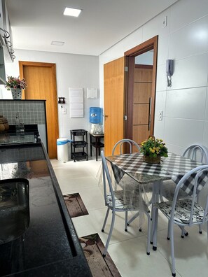 A modern kitchen with a black countertop, a water dispenser, and a dining area with a checkered tablecloth and metal chairs. - 3-bedroom apartment facing the sea on Morro Beach. (Guarapari)
