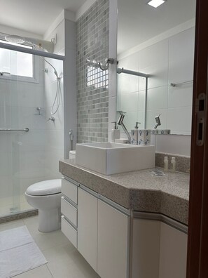A modern bathroom with a glass shower enclosure, a white toilet, a vanity with a mirror, and a granite countertop. - 3-bedroom apartment facing the sea on Morro Beach. (Guarapari)