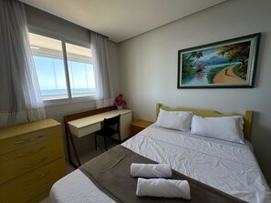 A hotel room with a bed, a desk, a chair, a dresser, and a painting on the wall. - 3-bedroom apartment facing the sea on Morro Beach. (Guarapari)