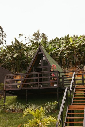 Une structure triangulaire en bois, dotée d’un balcon, et entourée d’une végétation luxuriante et de bananiers.