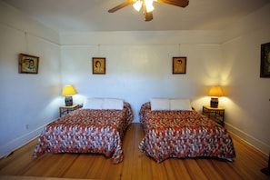 Una habitación con dos camas, piso de madera, ventilador de techo, lámparas de pared y cuadros enmarcados.