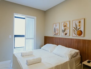 Ein Hotelzimmer mit Bett, Holz-Kopfstütze, Fenster mit Vorhängen und drei an der Wand befestigten Kunstwerken.