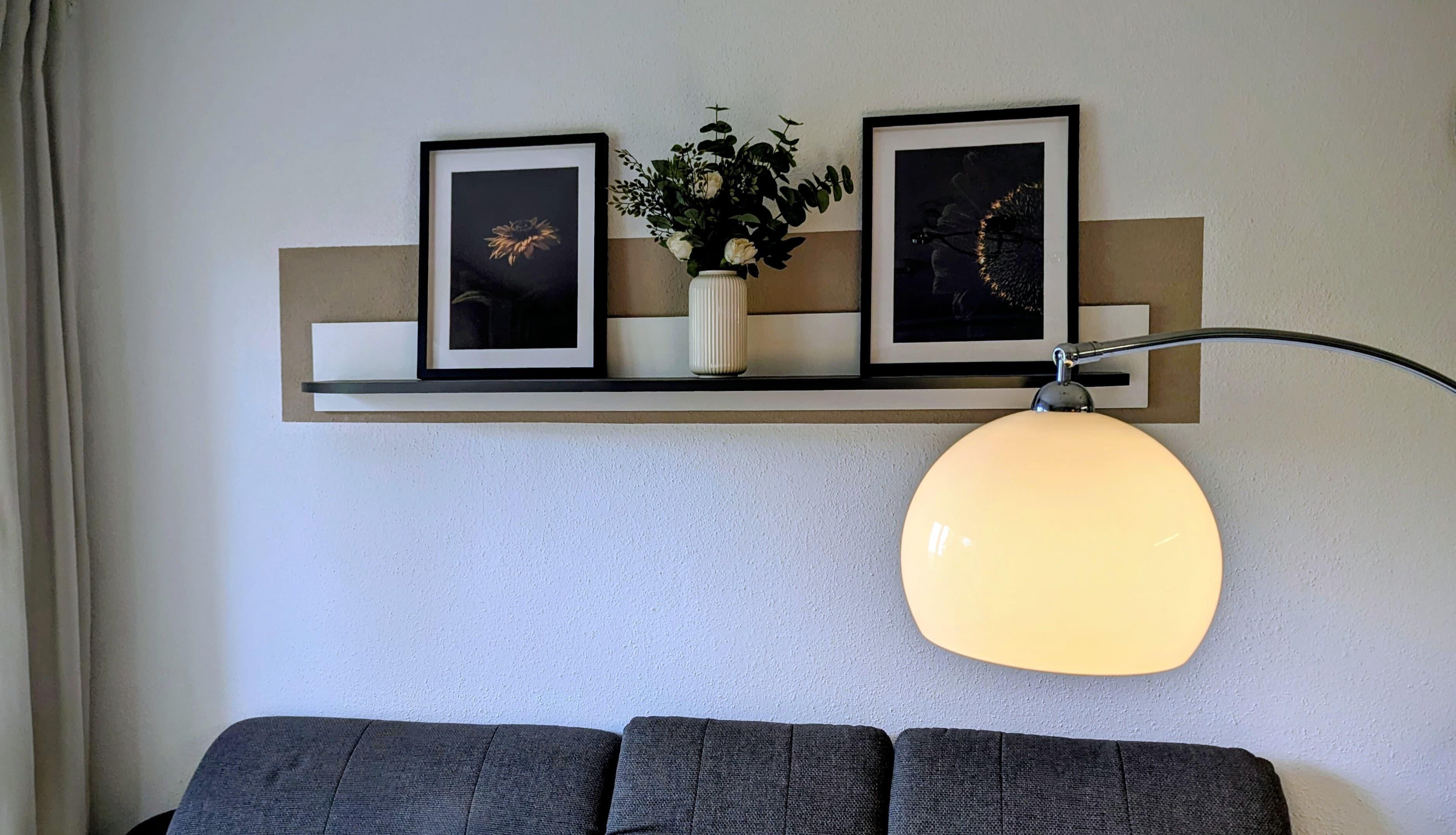 A living room with a sofa, a wall-mounted shelf with two framed pictures and a vase with flowers, and a floor lamp.