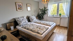 A double bed with floral bedding, a bedside table with a lamp, a window with curtains, and a radiator. - Vrbo Property (Schwarzenberg/Erzgebirge)