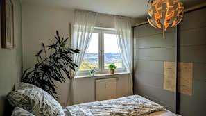 Ein Schlafzimmer mit großem Fenster, Heizkörper, Bett, Topfpflanze und einem einzigartigen Kronleuchter.