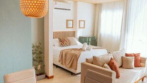 Quarto de hotel moderno com cama, sofá, mesa de jantar e uma xícara vermelha, além de luminária pendente.
